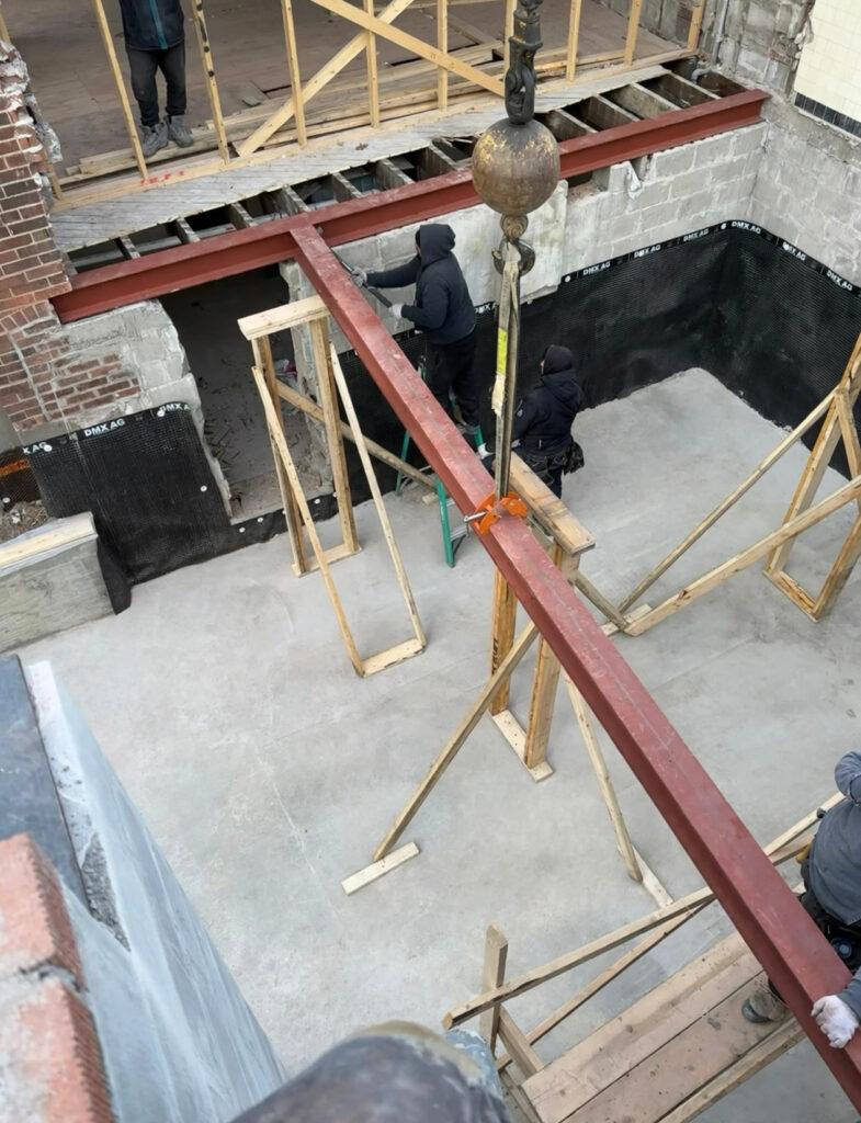 Steel beam being lowered into place by crane with temporary wooden supports during a structural renovation